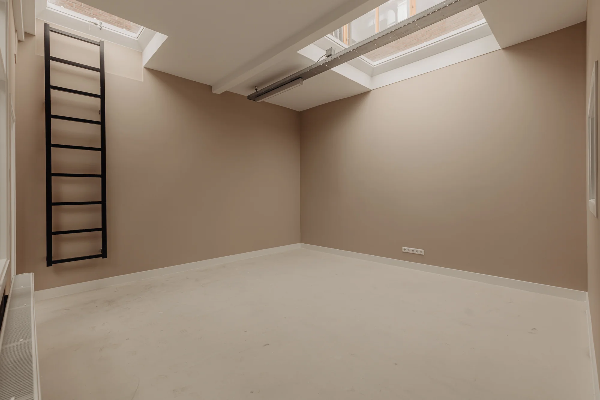 Lege kamer aan de Henri Polaklaan met beige muren, dakramen en een zwarte ladder tegen de muur.